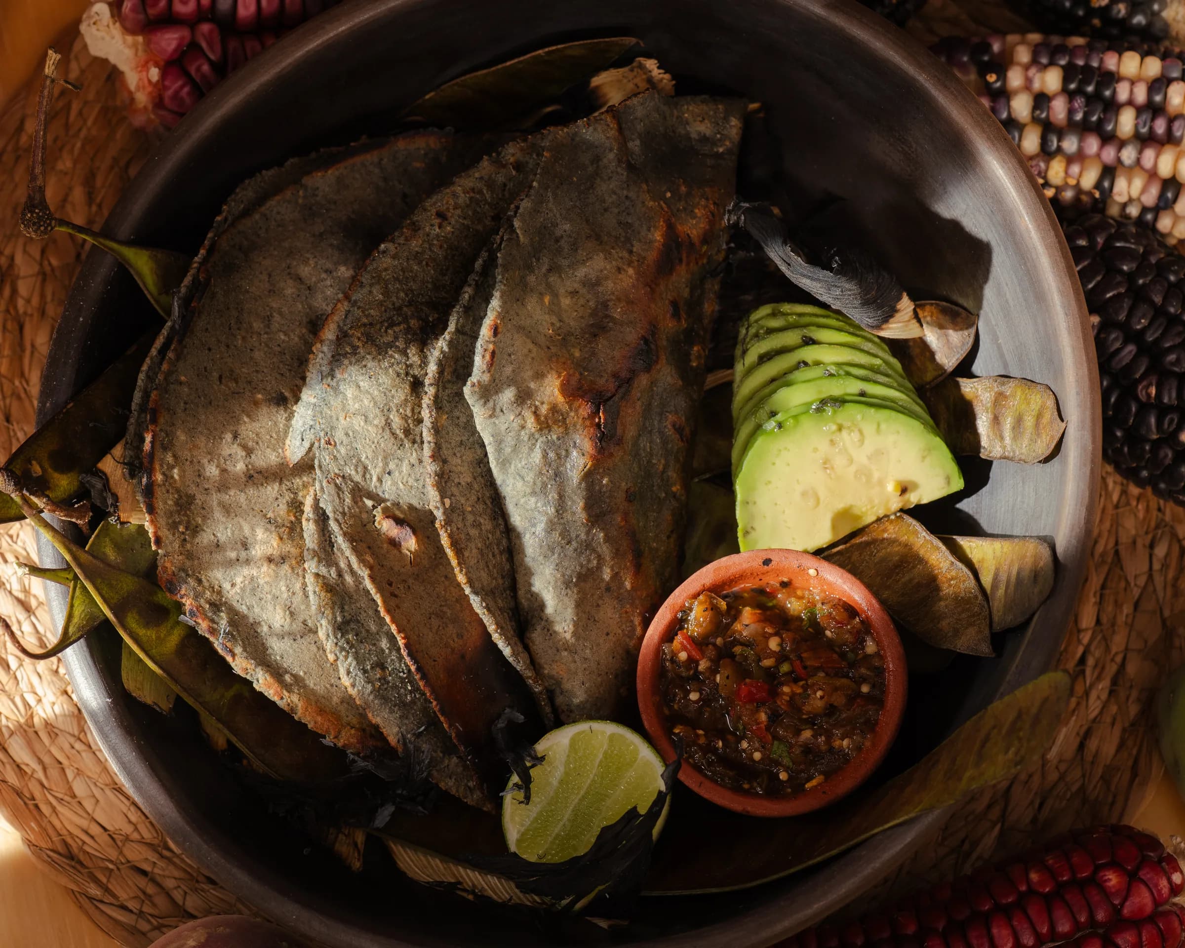 Tortillas oscuras con aguacate y salsas sobre un plato negro.