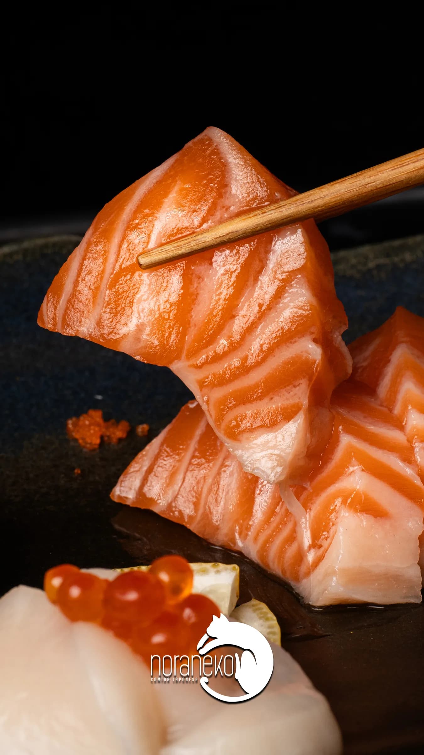 Palillos sosteniendo una pieza de salmón sobre un plato oscuro.