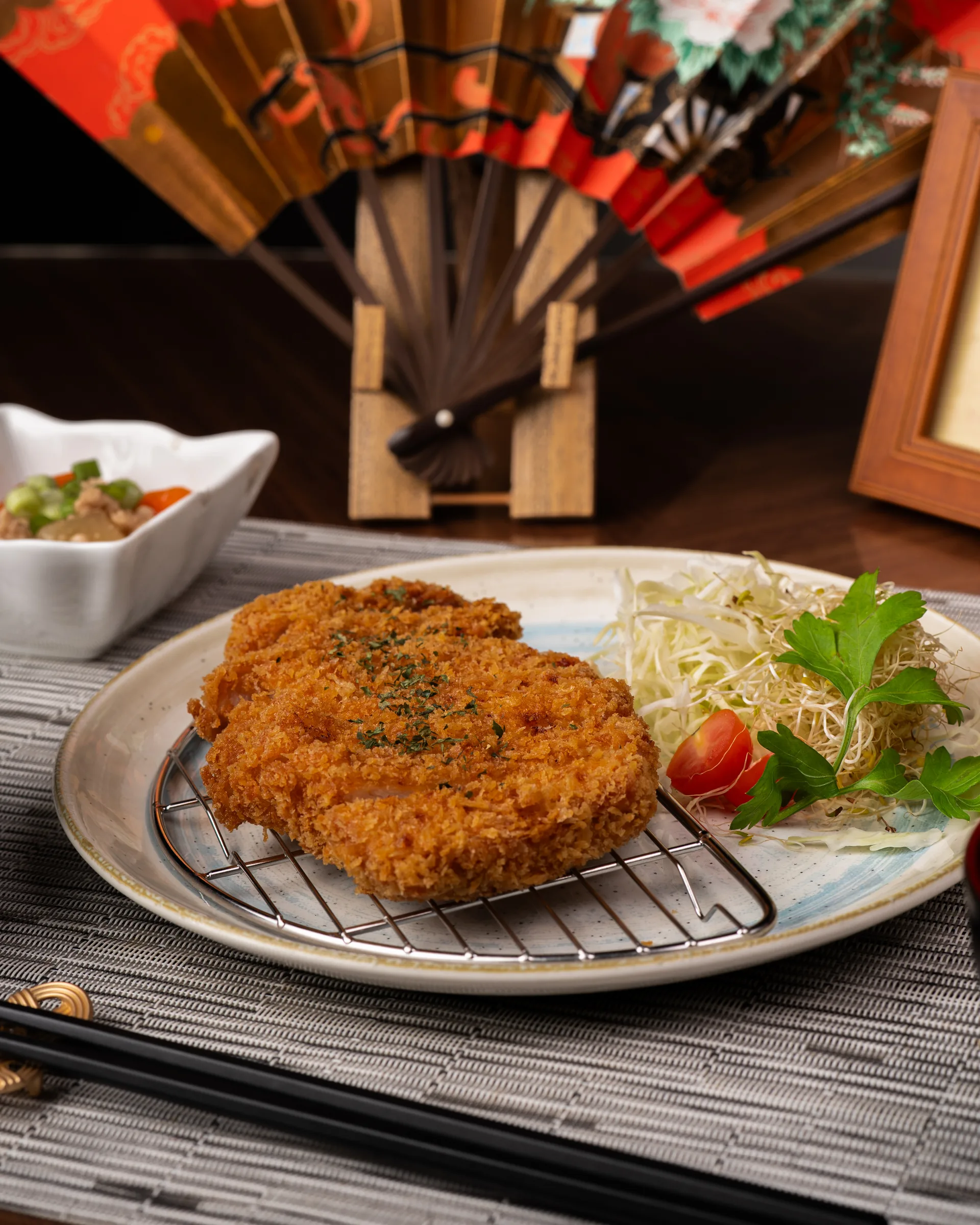Plato empanizado con guarnición y elementos de mesa al fondo.